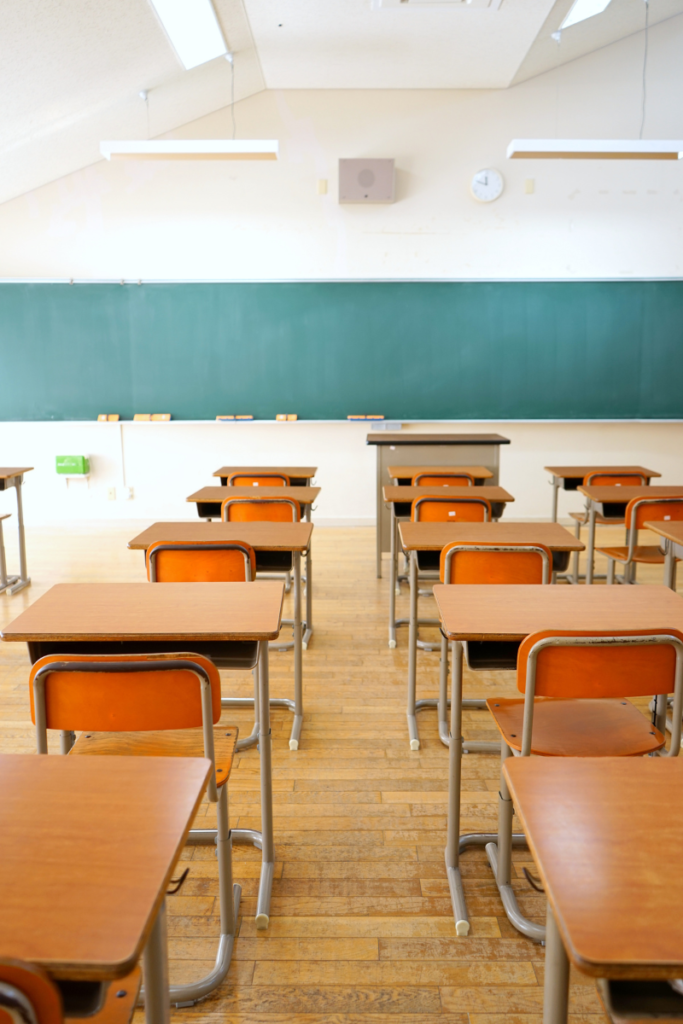 A classroom with rows of wooden desks and orange chairs and a large blackboard on the wall