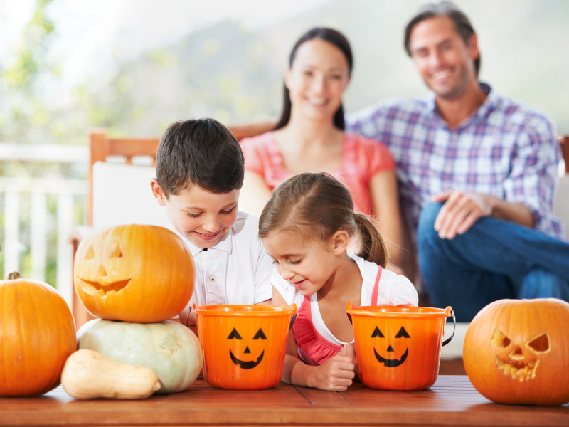 Two kids looking into pumpkin Halloween baskets with parents sitting in the background