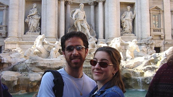 A young couple standing in front of the Trevi Fountain on a sunny day.
