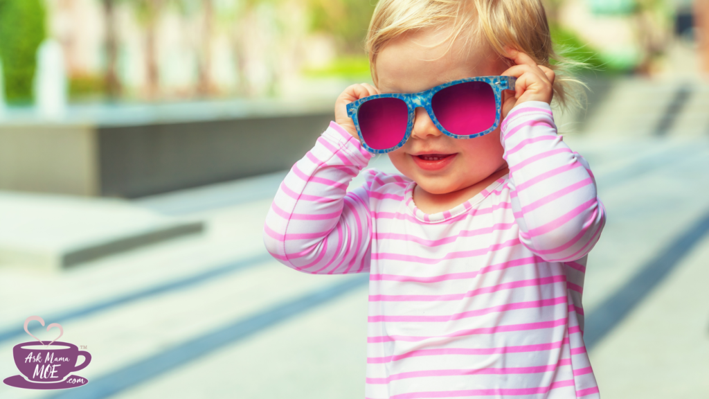 A toddler girl, wearing a white and pink striped shirt and wearing a pair of sunglasses