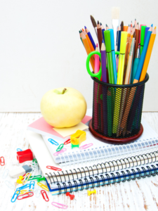 Pencil holder filled with pens, pencils, scissors, sitting on a stack of books with an apple beside it.