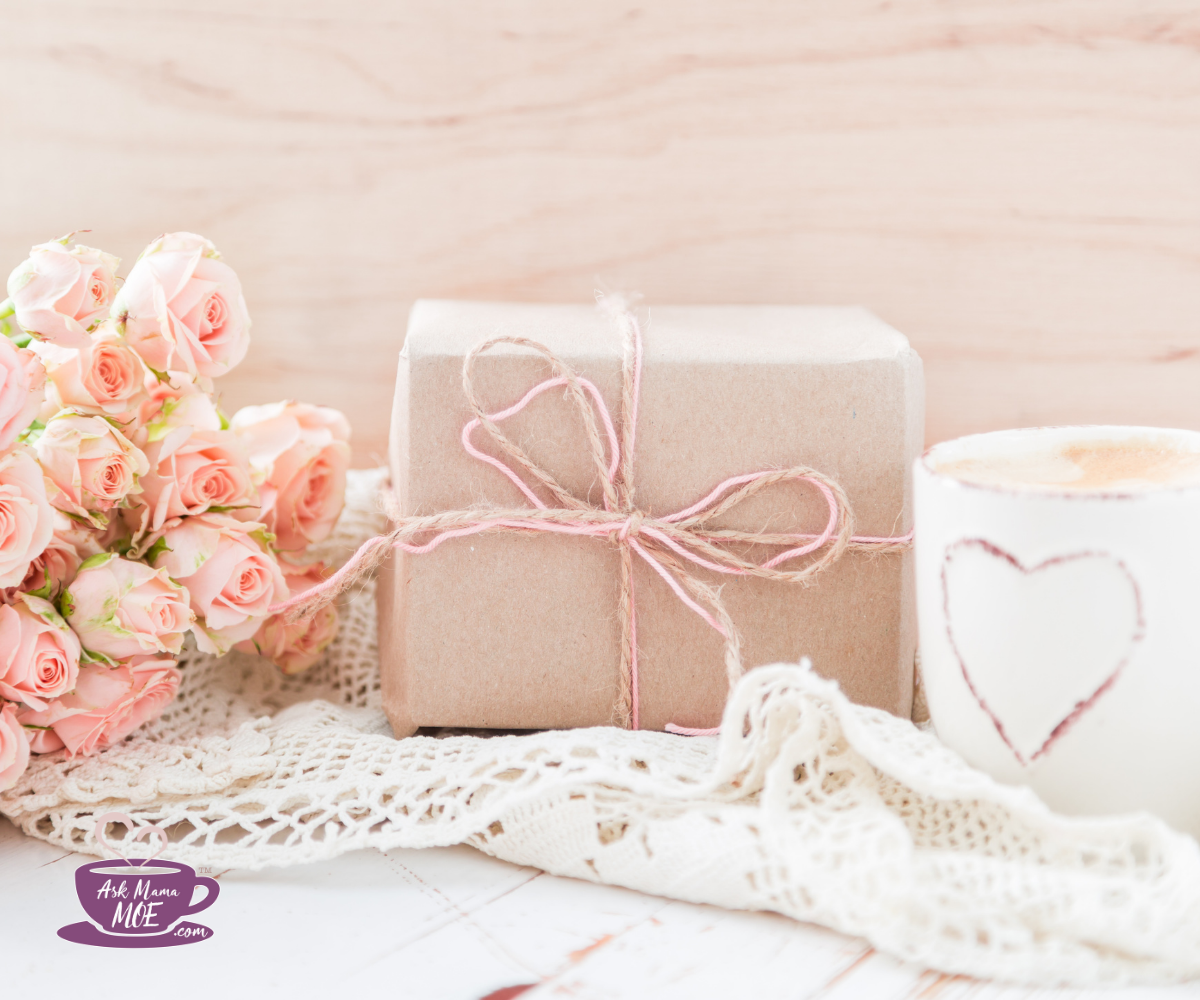 A gift wrapped in light brown kraft paper and tied with pink ribbon, surrounded by roses and a mug.